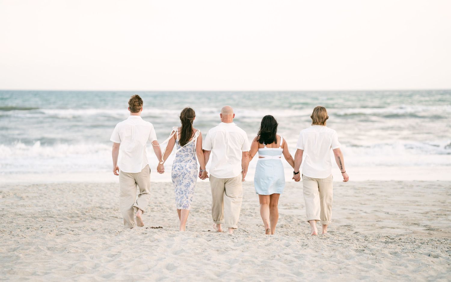 Family of five holding hands walking toward ocean on sandy beach, viewed from behind.