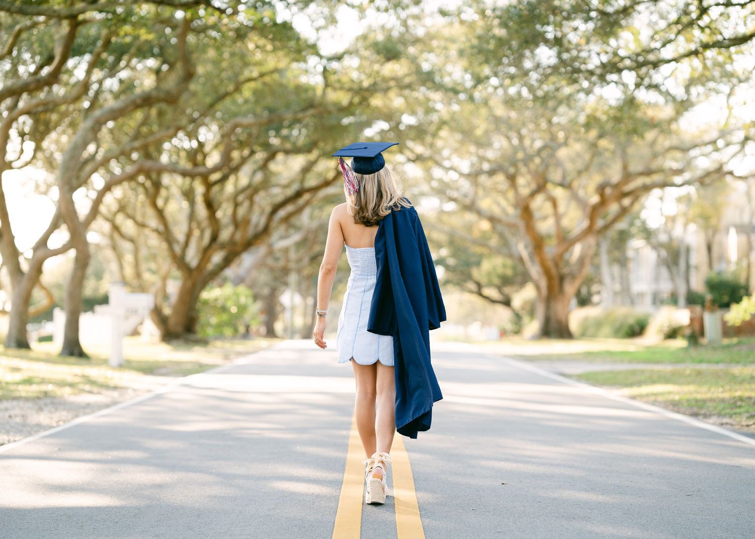 Graduate walking down tree-lined road holding navy cap and gown in white dress.