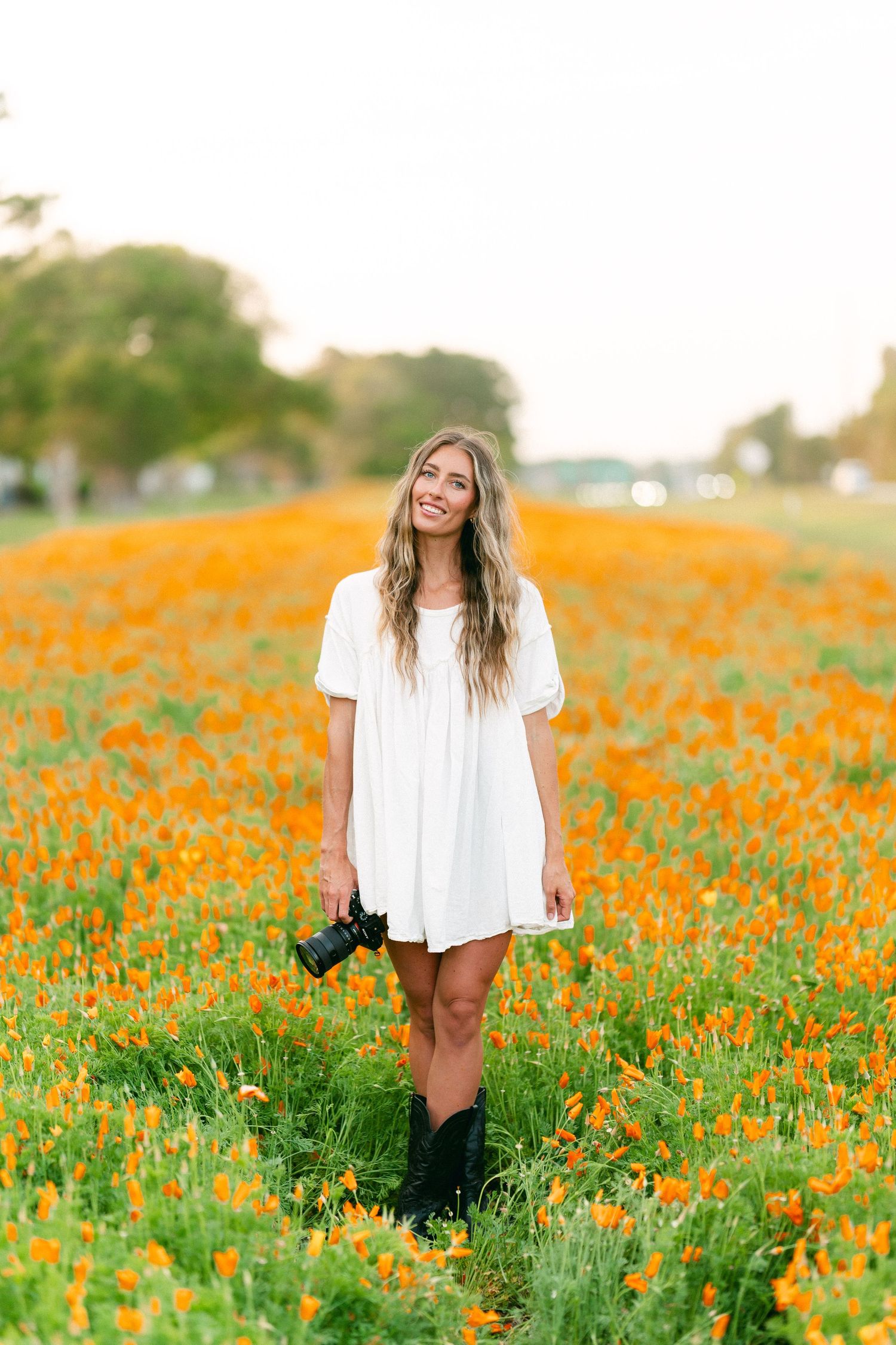 Woman in white dress holding camera standing in vibrant orange poppy field wearing black boots.