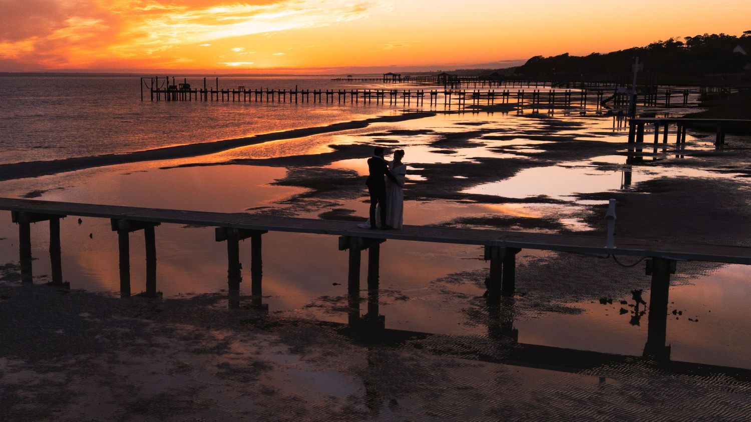 Dramatic sunset silhouettes an old weathered pier reflecting warm orange hues over calm coastal waters.