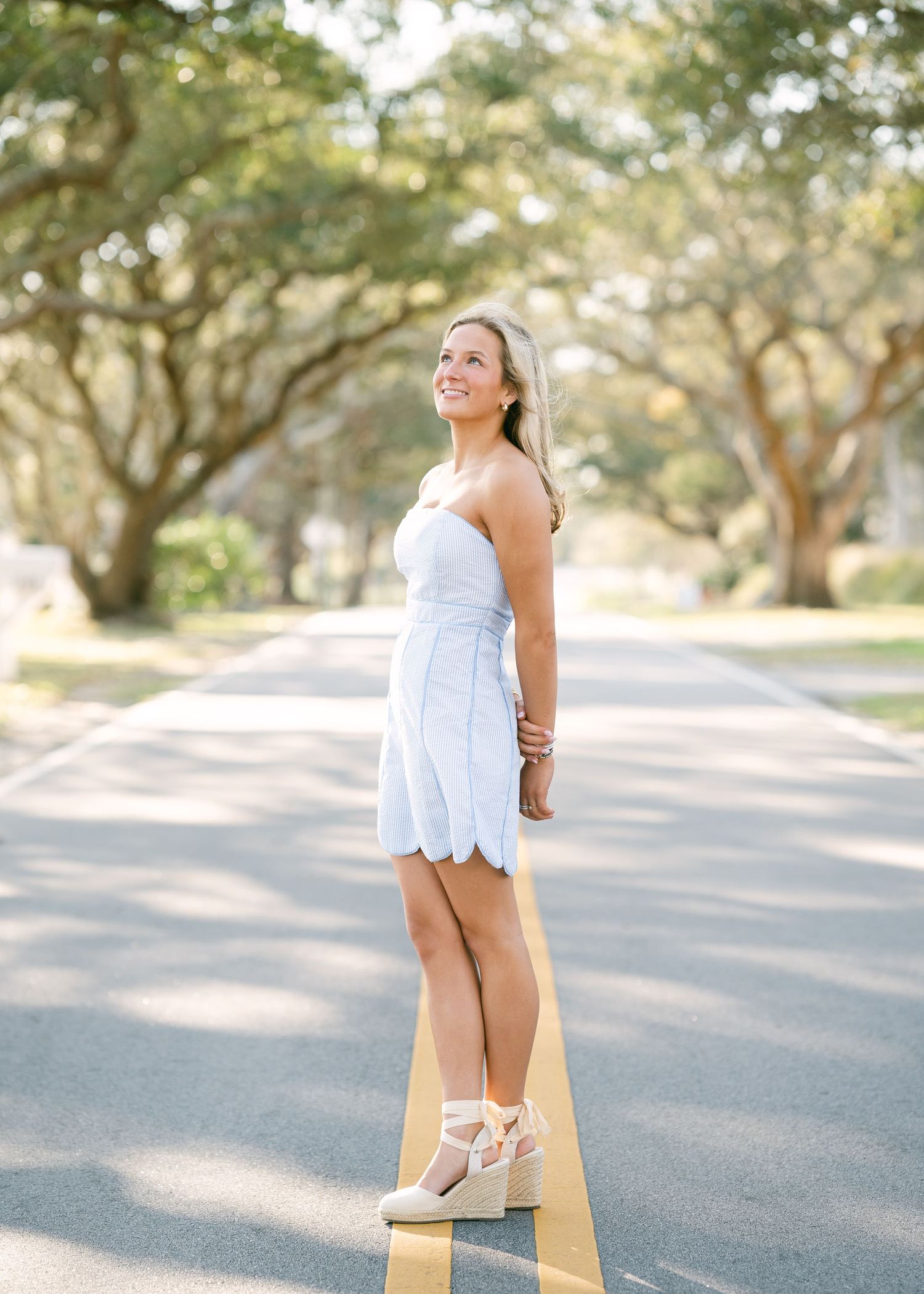 Blonde woman in white scalloped mini dress standing on tree-lined road during senior portrait.