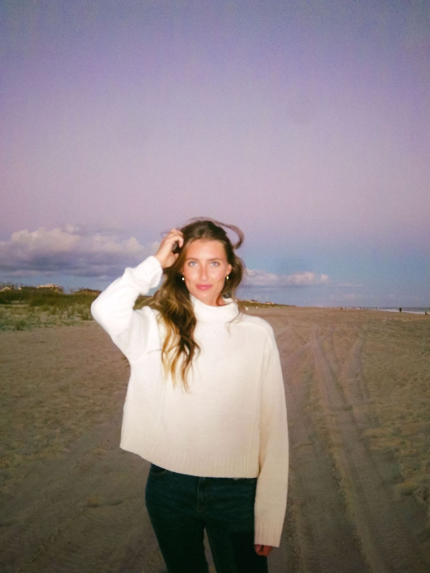 Young woman in white turtleneck sweater standing on a beach at dusk with purple sky behind her.