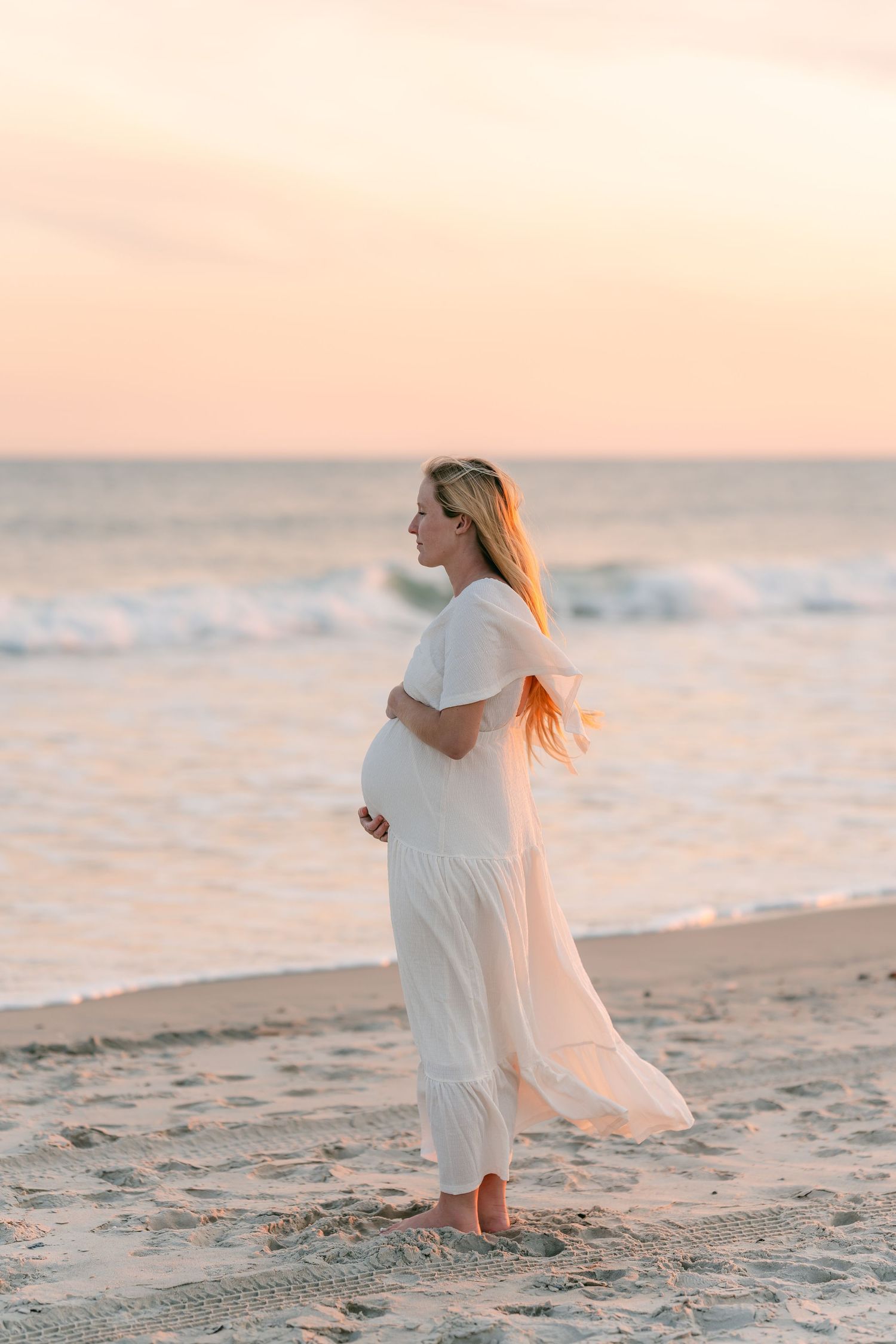 Pregnant woman in white dress standing on beach at golden sunset, cradling baby bump near ocean waves.