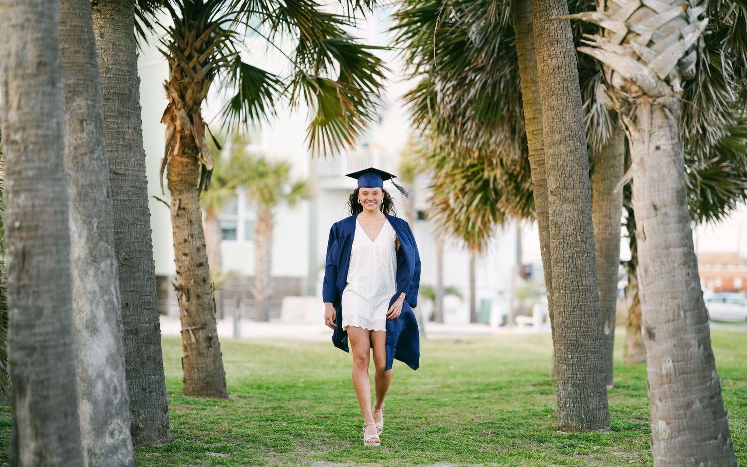 Graduate in navy cap and gown walking between palm trees on green campus lawn.
