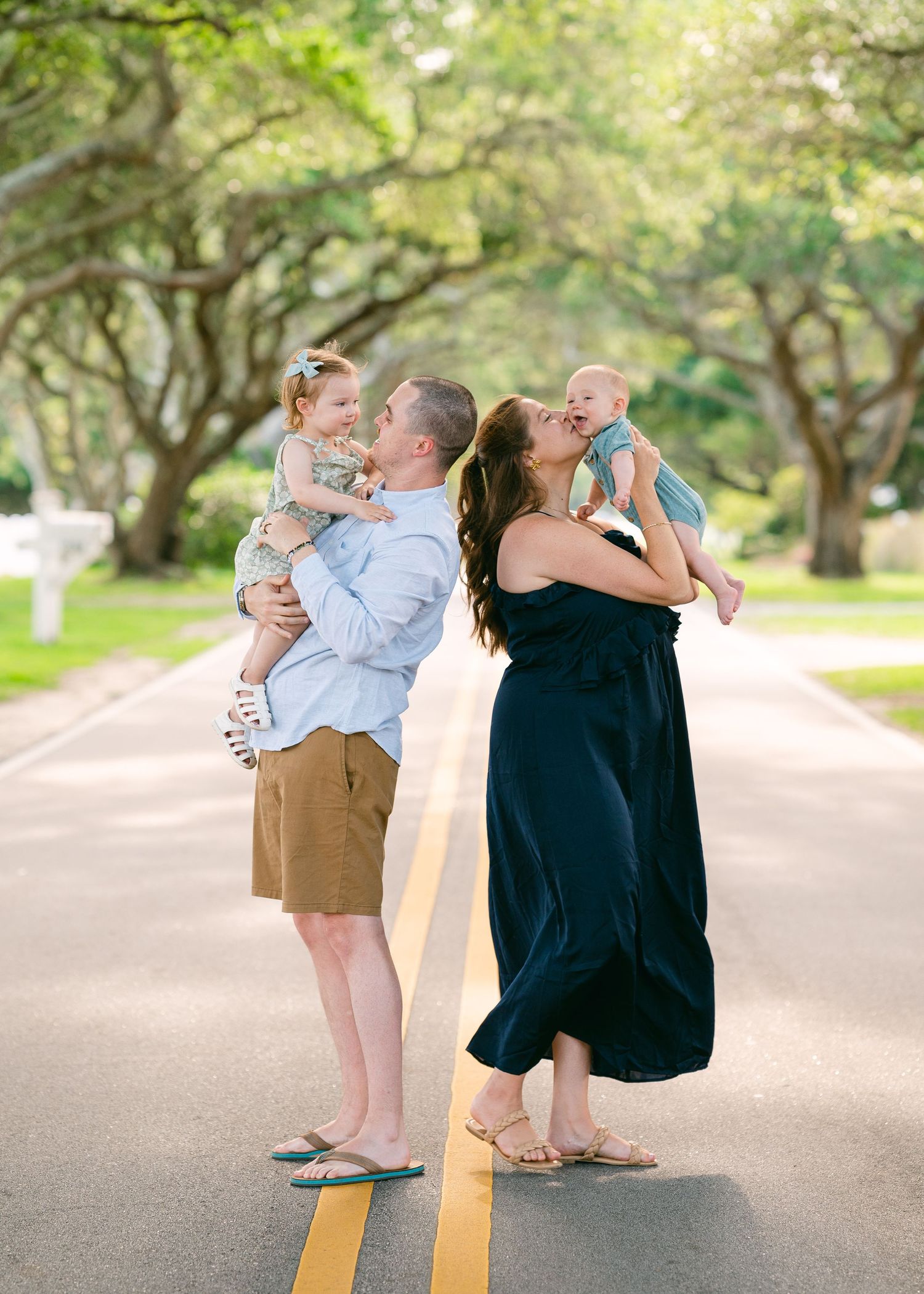 Couple holding two babies on tree-lined road in warm outdoor family portrait session.