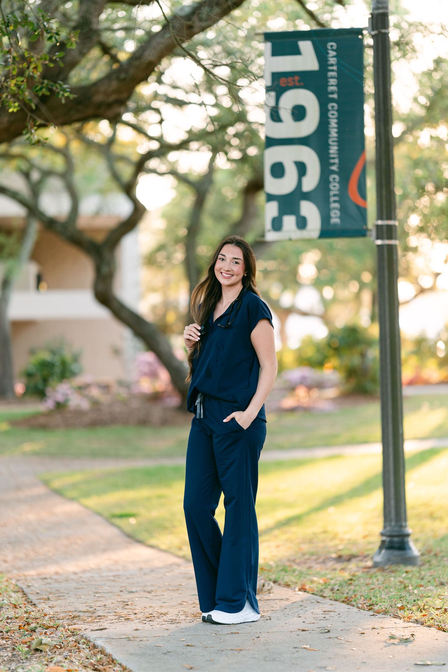 Woman in navy scrubs smiling beside Carteret Community College 1963 banner on campus walkway.