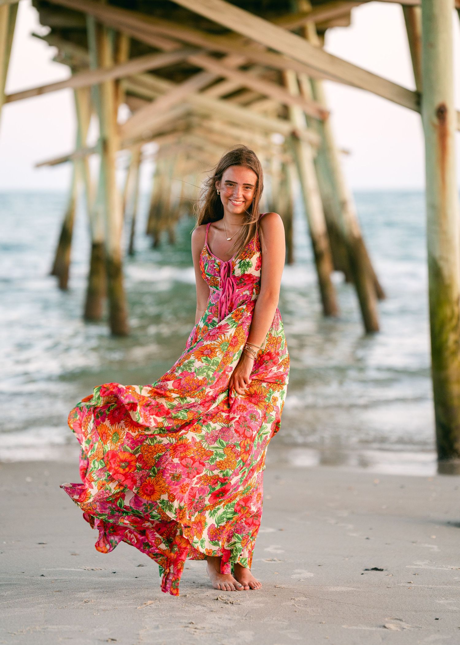 Young woman in vibrant floral maxi dress posing gracefully under a wooden beach pier.