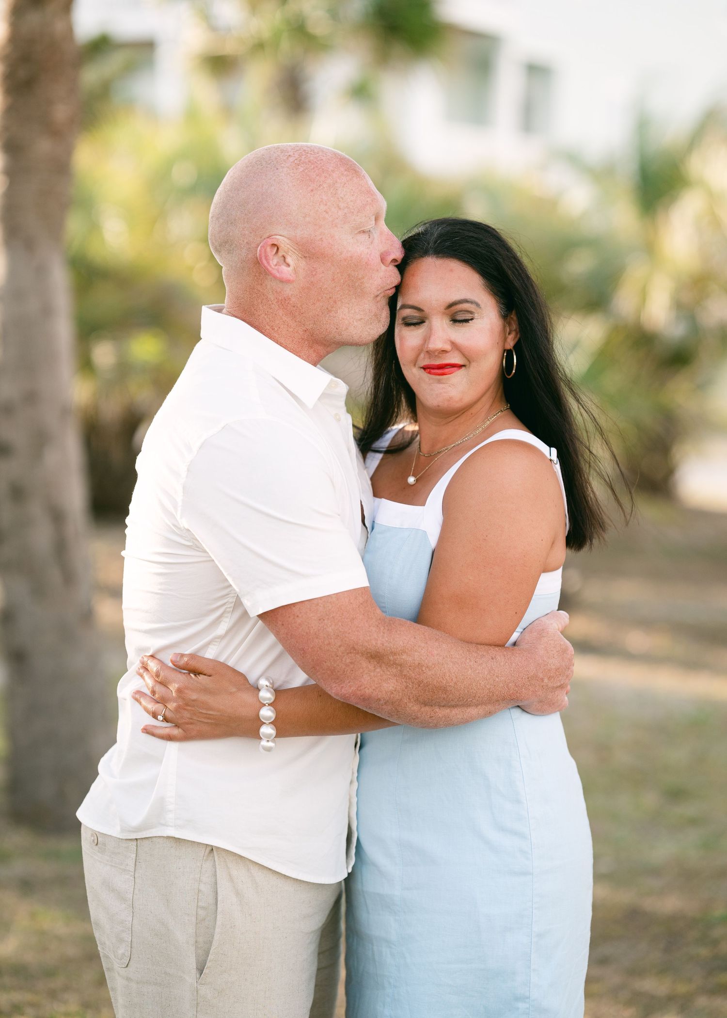 Bald man kissing dark-haired woman on cheek outdoors, both smiling, surrounded by tropical greenery.
