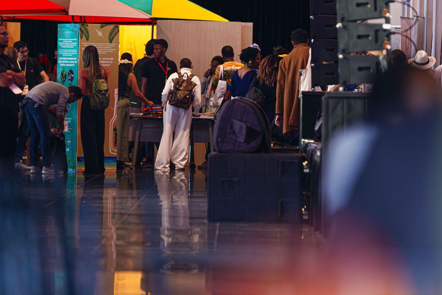 People browsing vendor booths at an indoor market or convention hall with colorful umbrellas and display tables.