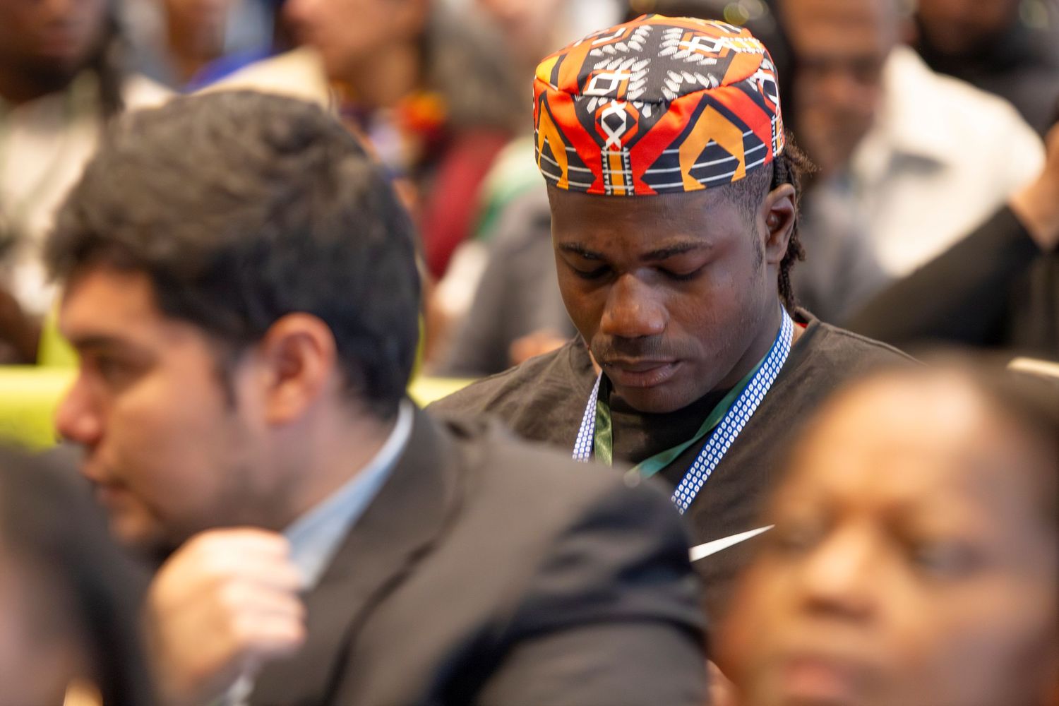 Diverse attendees in formal attire at a conference, man in colorful African kufi cap looking down thoughtfully.