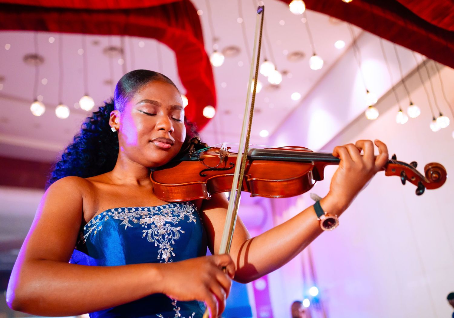Musicians performing with violins under pink and purple stage lighting.