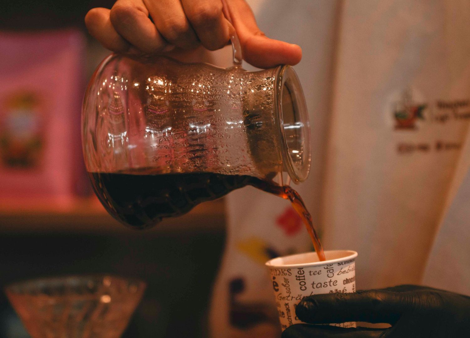 Barista pouring rich dark coffee from a glass carafe into a paper cup at a specialty coffee shop.