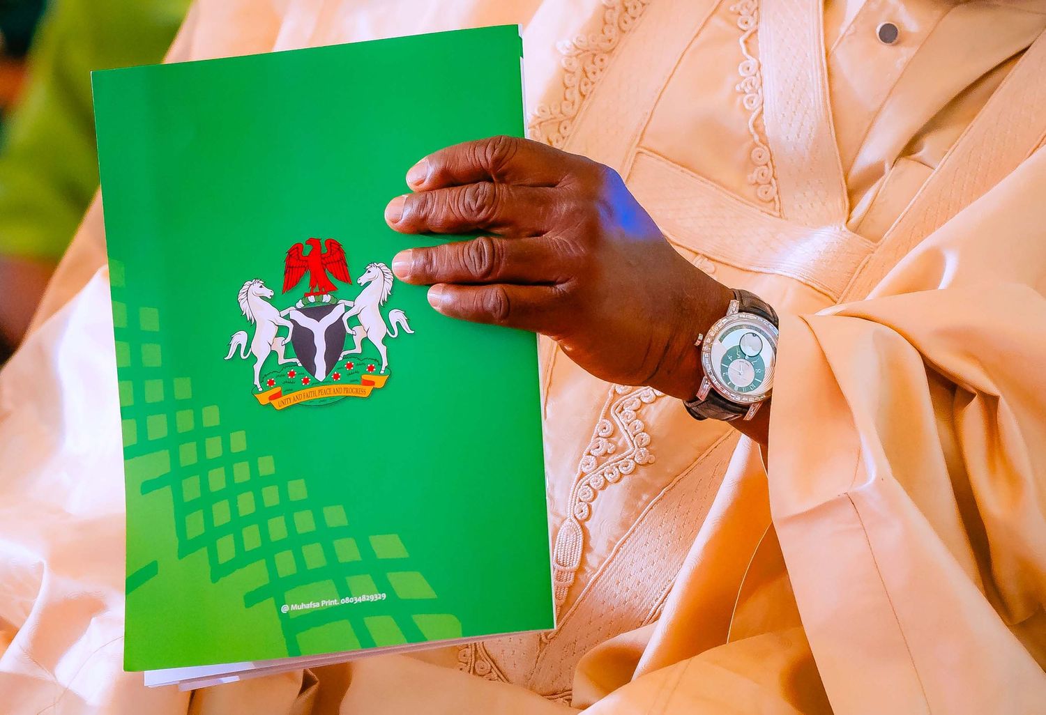 A person holds a green Nigerian passport against a white background while wearing traditional attire.