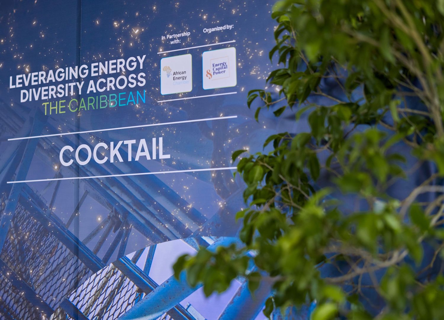 Sign for a Caribbean energy diversity cocktail event displayed outdoors with tropical foliage in the foreground.