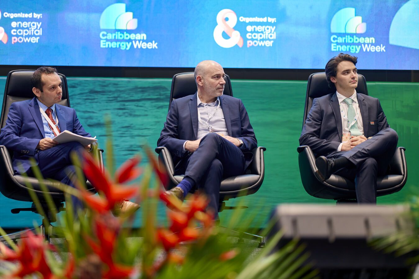 Three men in suits seated on stage at Caribbean Energy Week conference panel discussion.