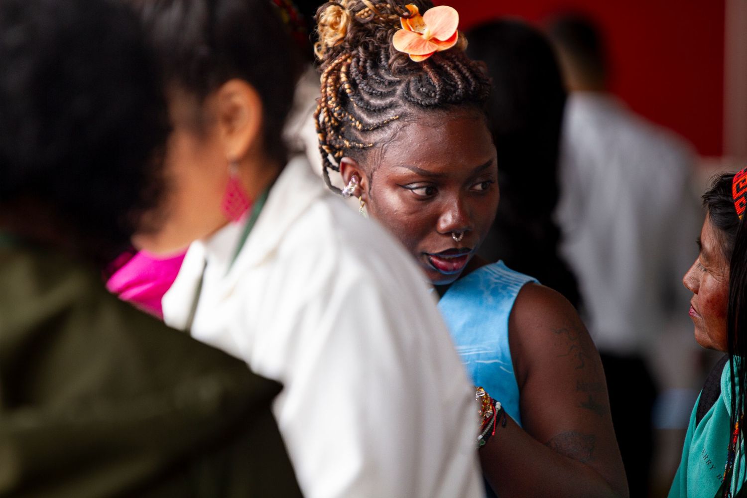 A young African woman with braided hair and orange flower accessory stands among a group of people indoors.