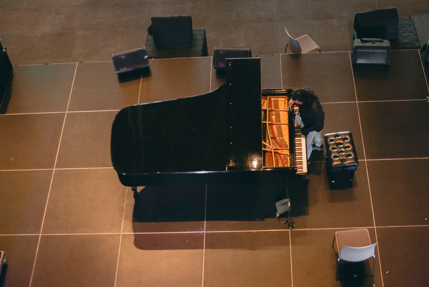 Aerial view of a black grand piano being played in a spacious hall with brown tiled flooring.