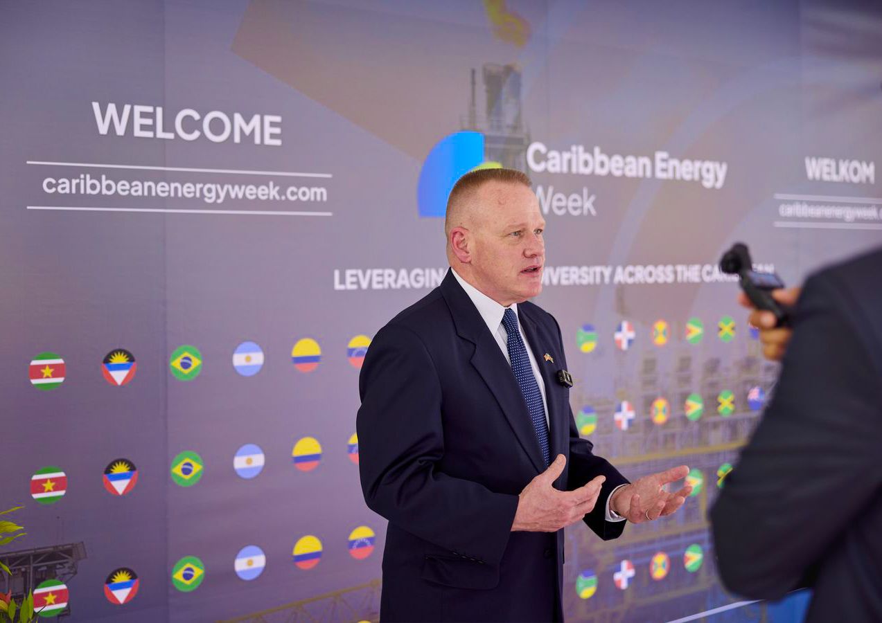 Man in suit speaking at Caribbean Energy Week conference with welcome banner and country flags backdrop.