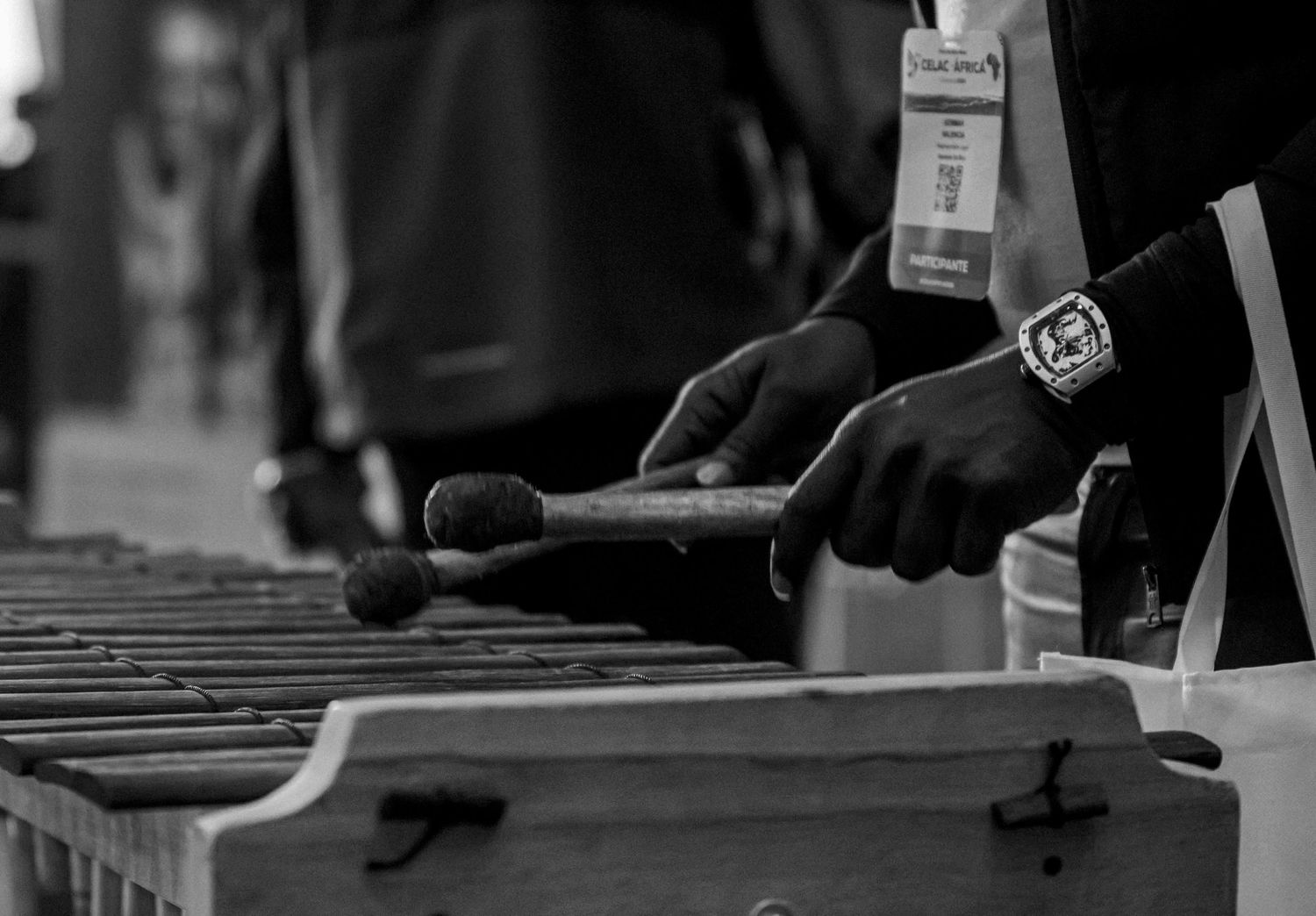 Black and white photo of hands playing a marimba with mallets, wearing a watch, at a live music event.