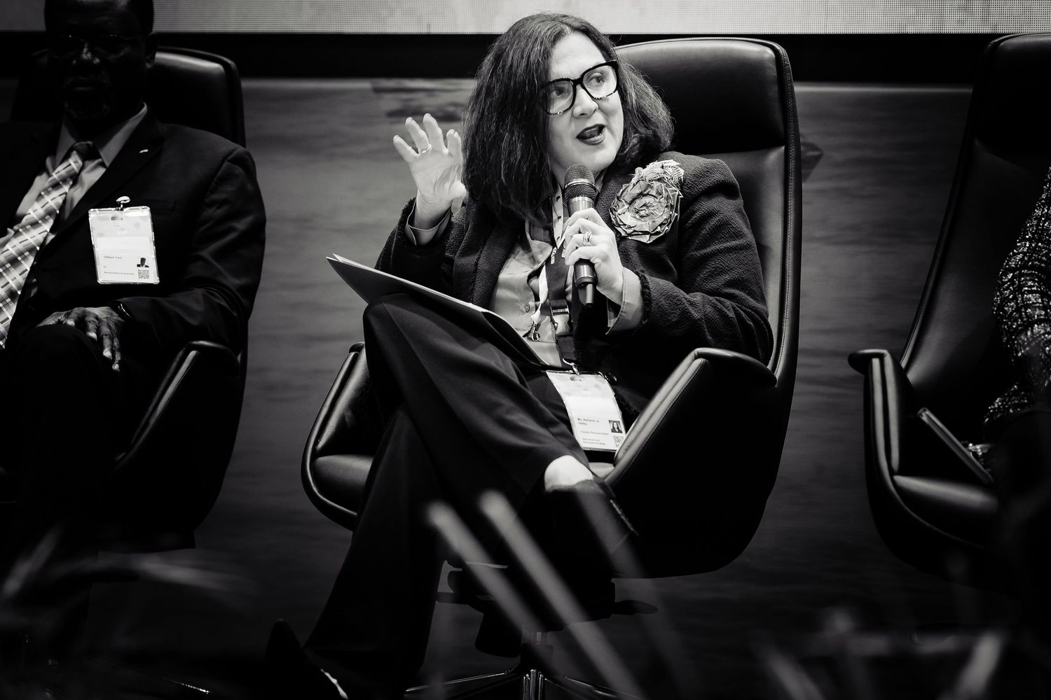 Woman speaking at conference panel, holding microphone, seated in black chair, black and white professional event photo.