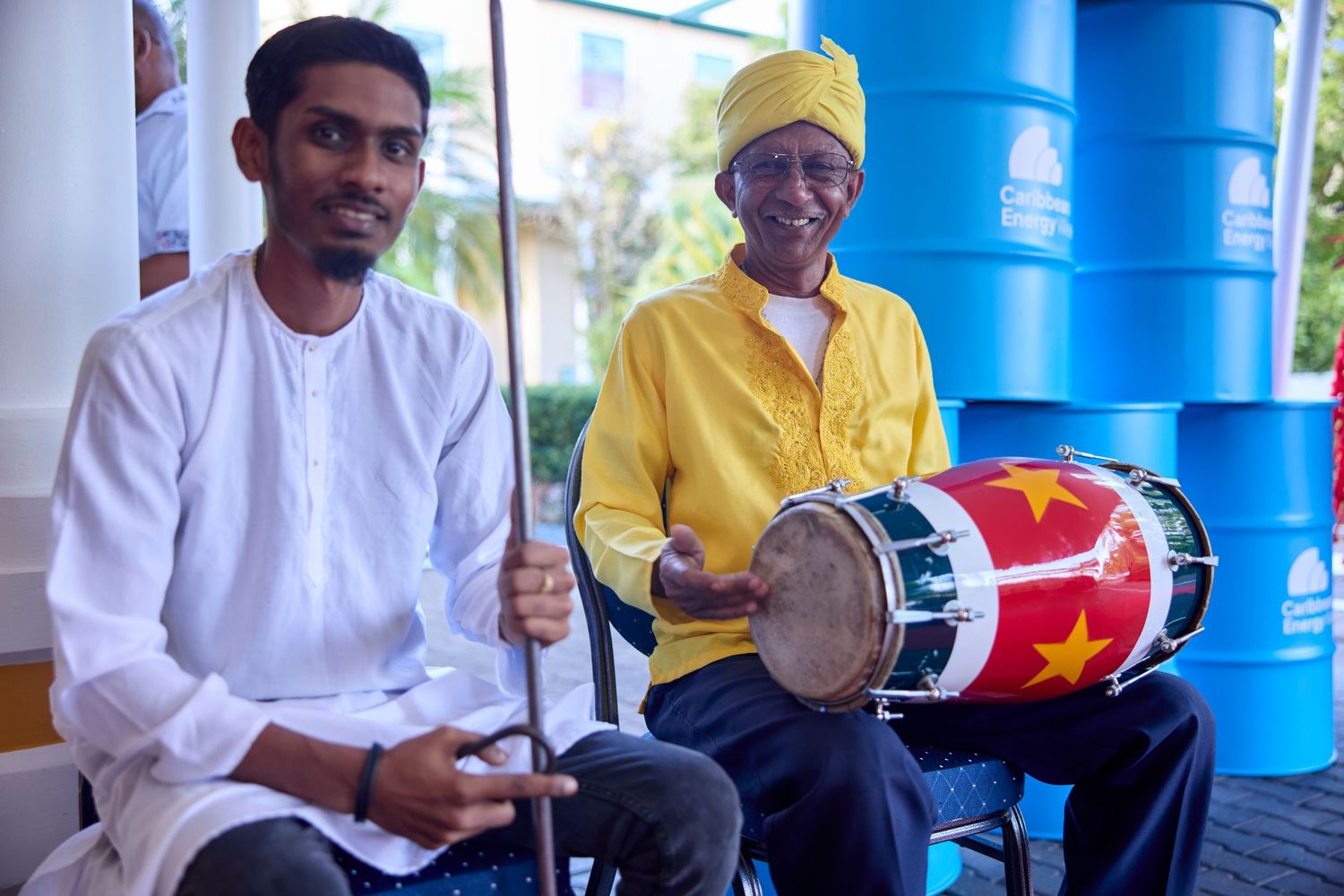 Two musicians in traditional attire, one in white and one in yellow turban, playing drums outdoors.