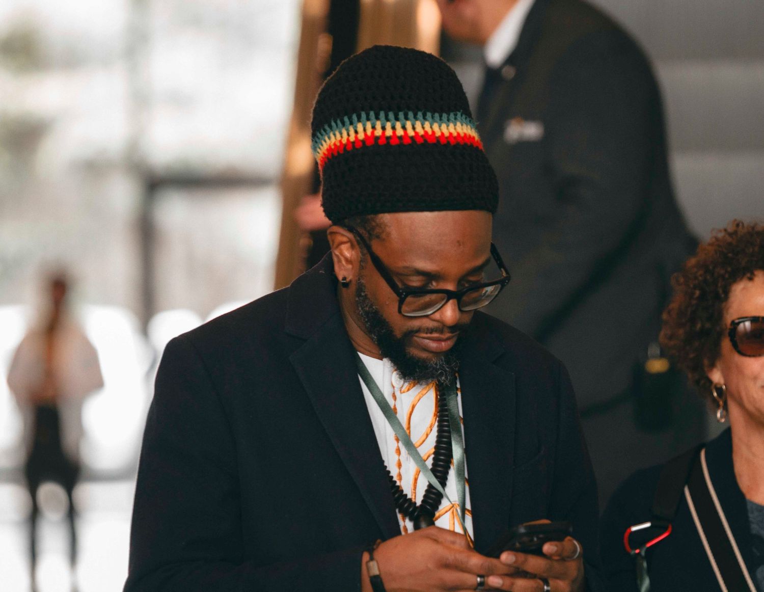 Man in black suit and colorful knit hat looks at smartphone while wearing lanyard at indoor event.