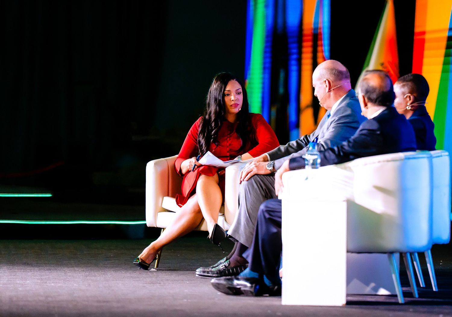 People seated on a brightly lit stage with colorful vertical light beams in the background during a conference panel.