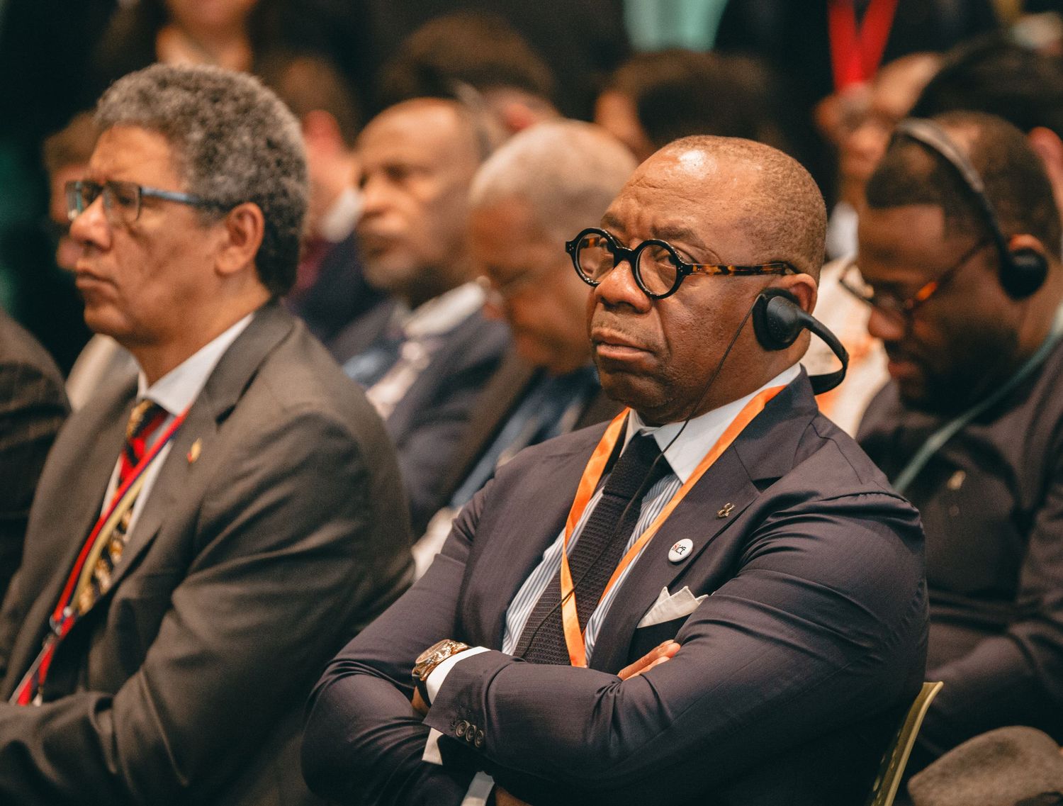 Business professionals in suits with orange lanyards seated at a formal conference or summit event.