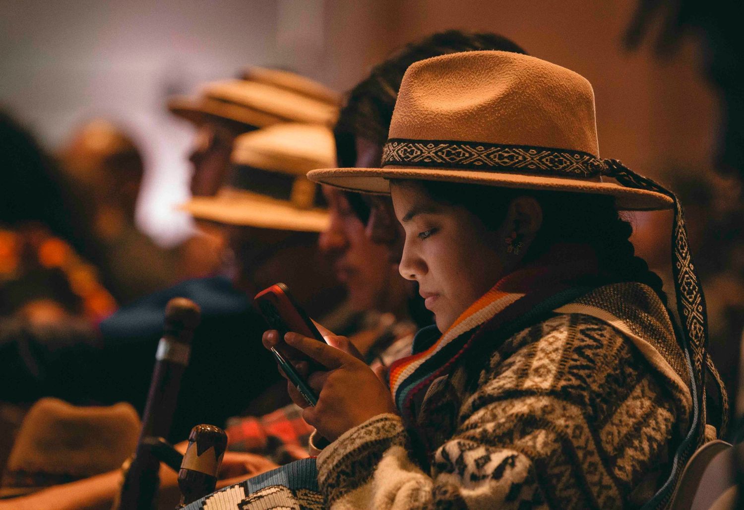 Young child in tan cowboy hat and patterned sweater focuses on smartphone in dimly lit room.