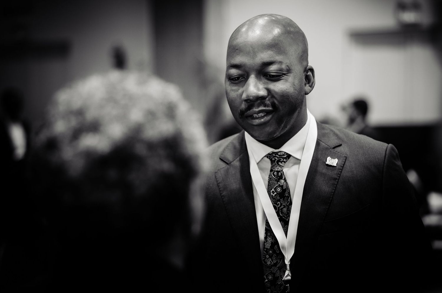 Black and white photo of a smiling man in a suit and lanyard at a professional event.