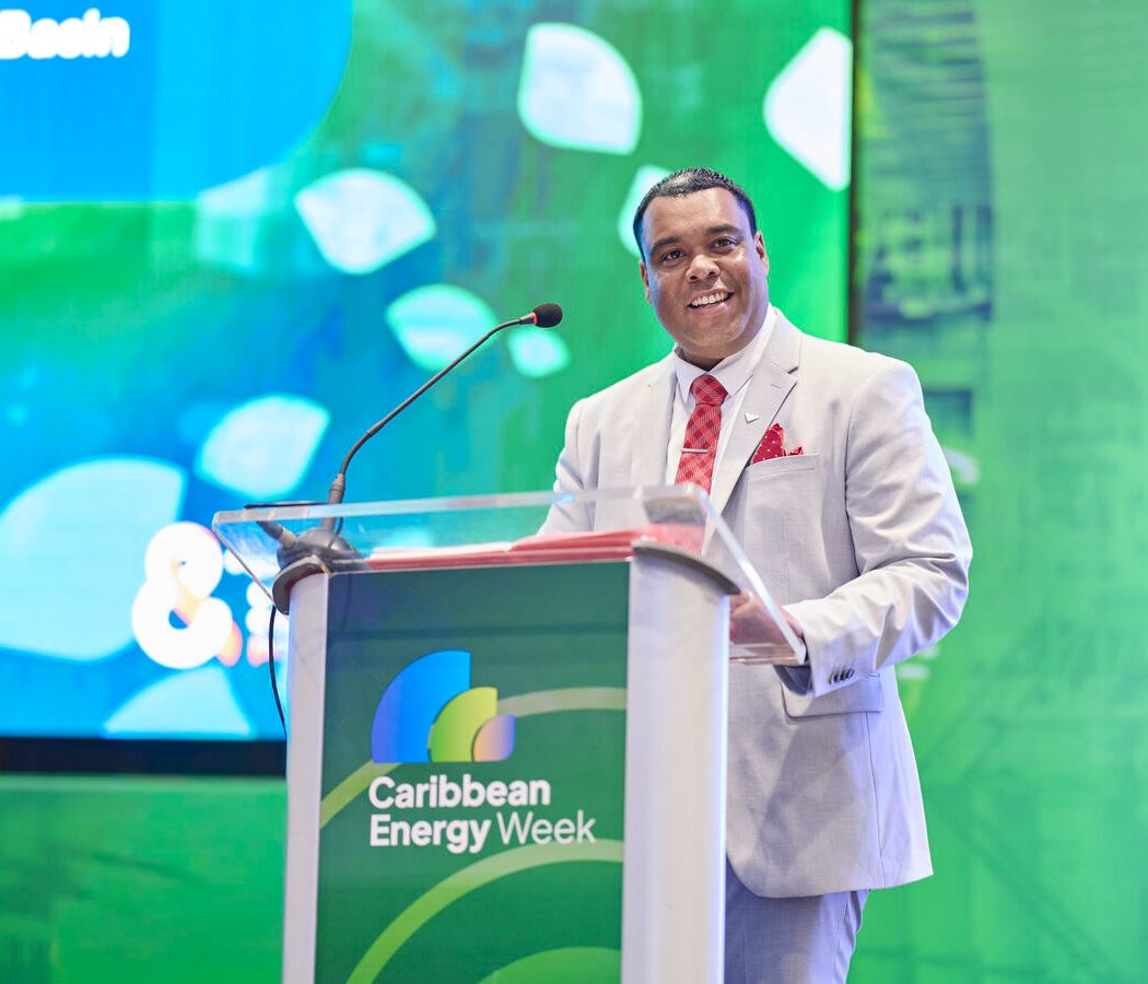 Man in white suit speaking at Caribbean Energy Week conference podium with green background.