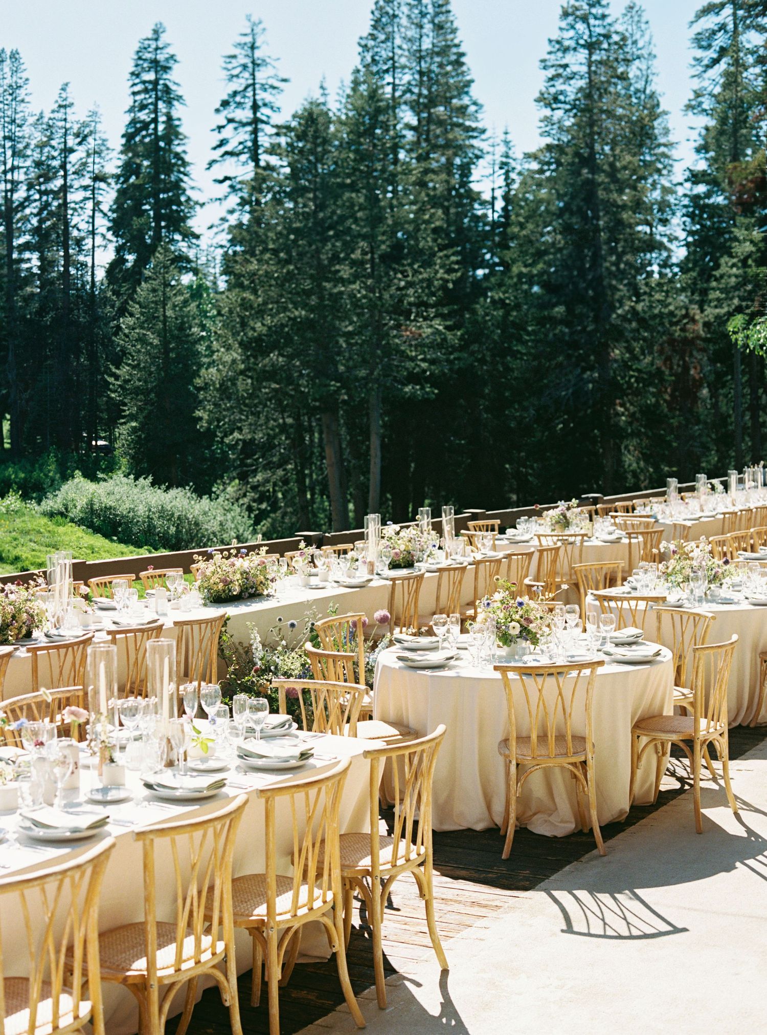An elegant outdoor wedding reception setup with wooden chairs and round tables against a forest backdrop.
