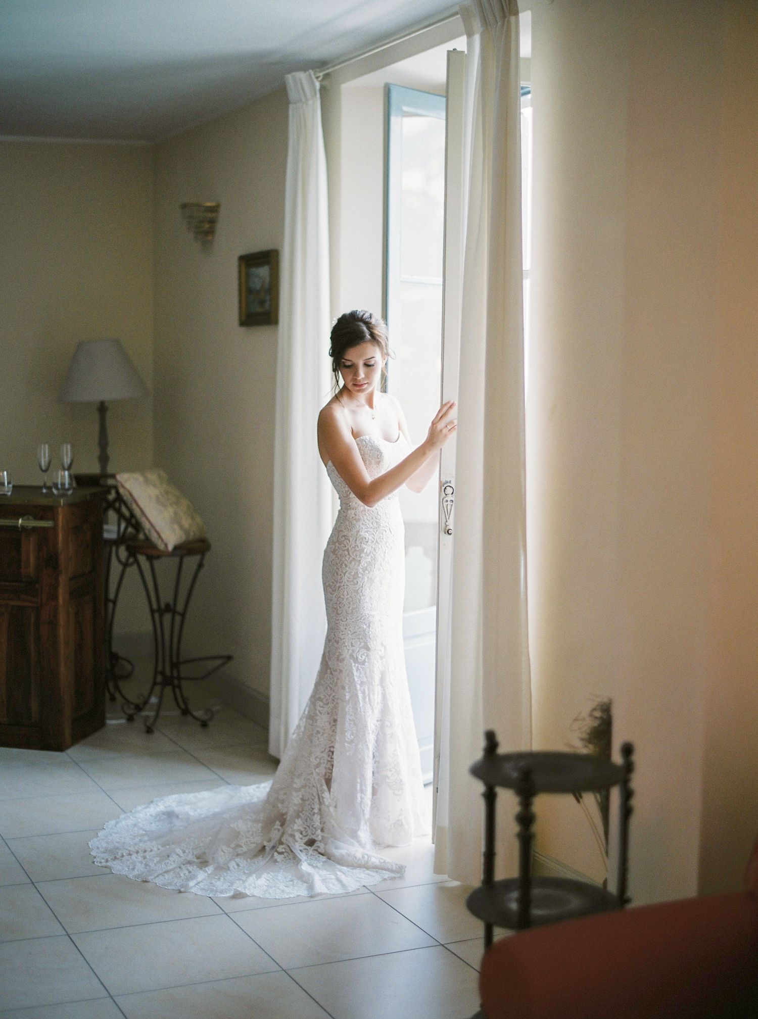 A white lace wedding dress is showcased in a bright indoor room with natural window light.