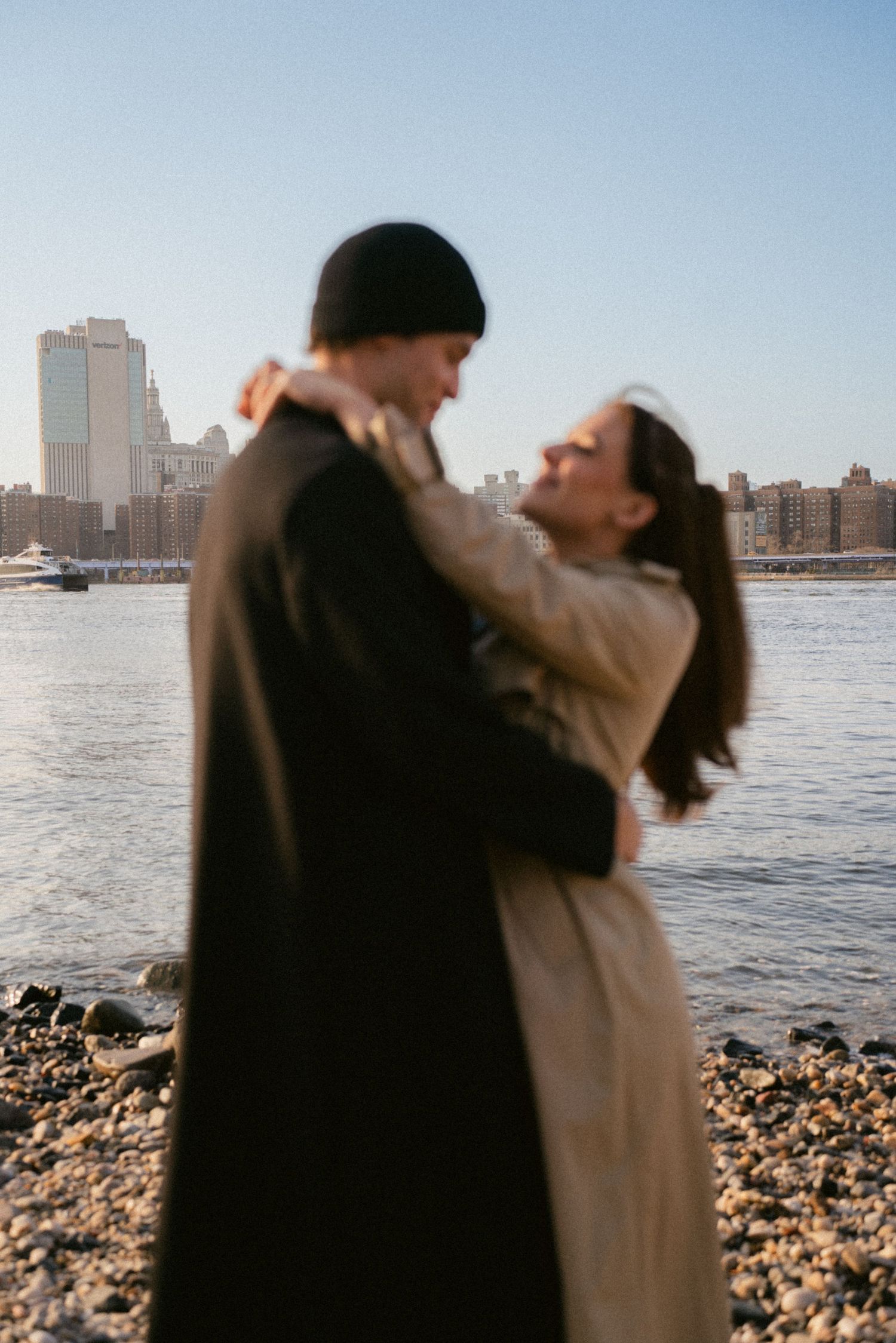 Romantic couple poses playfully by the waterfront with city skyline in background during sunset.