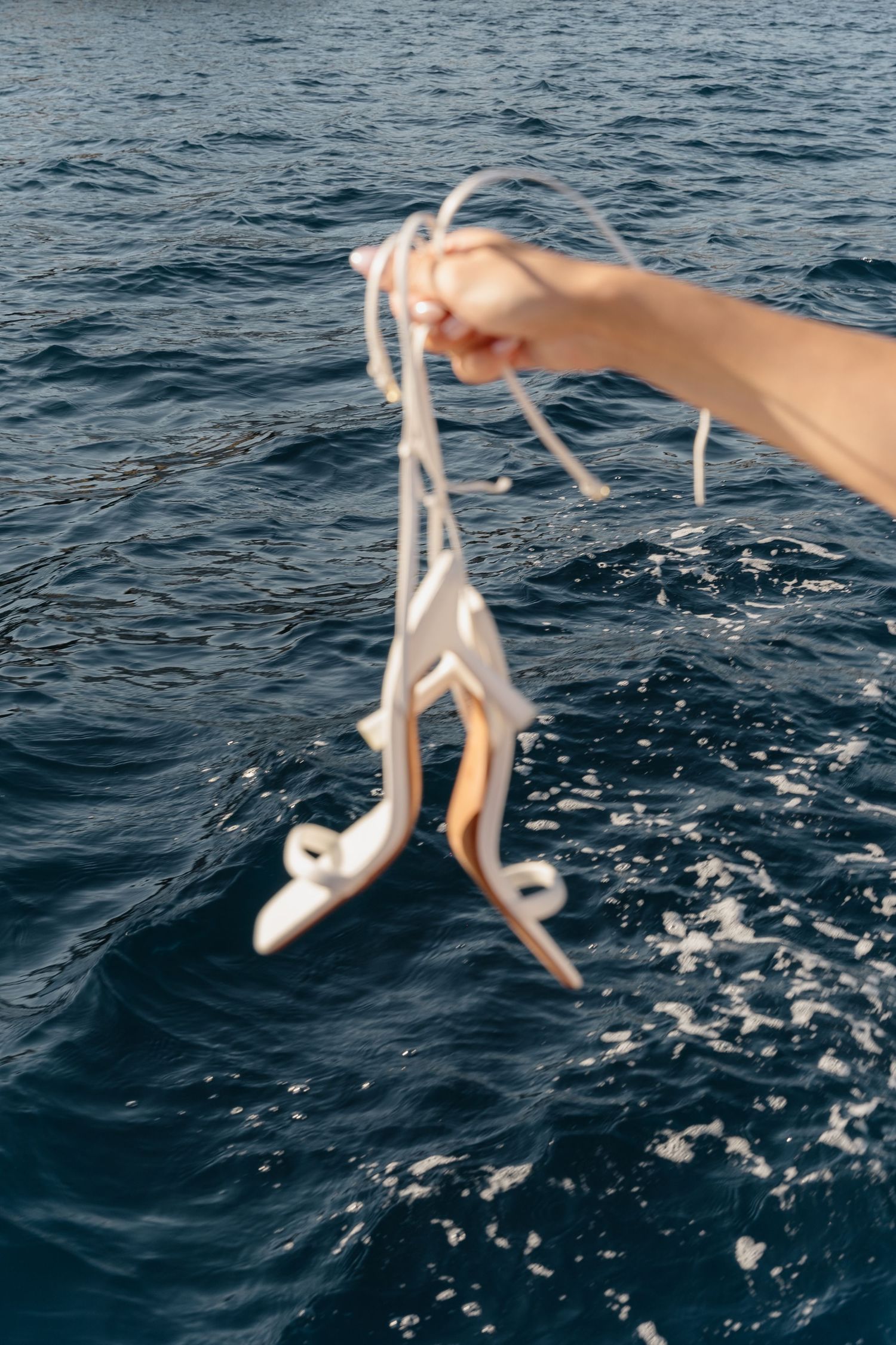 Hand holding a pair of white strappy sandals by their laces over dark blue ocean water with small splashing waves.