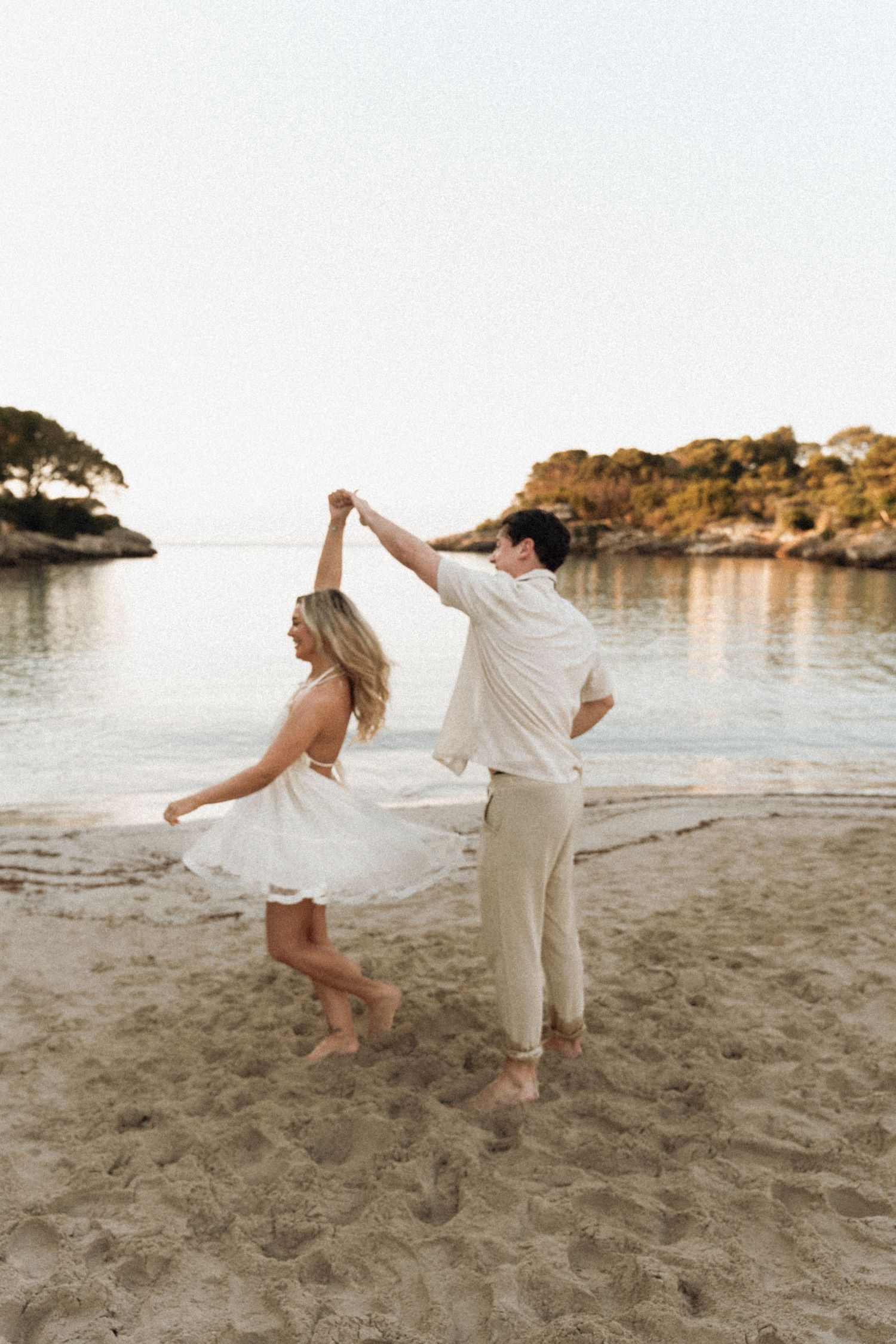 A couple in white clothing dances playfully on a sandy beach at sunset.