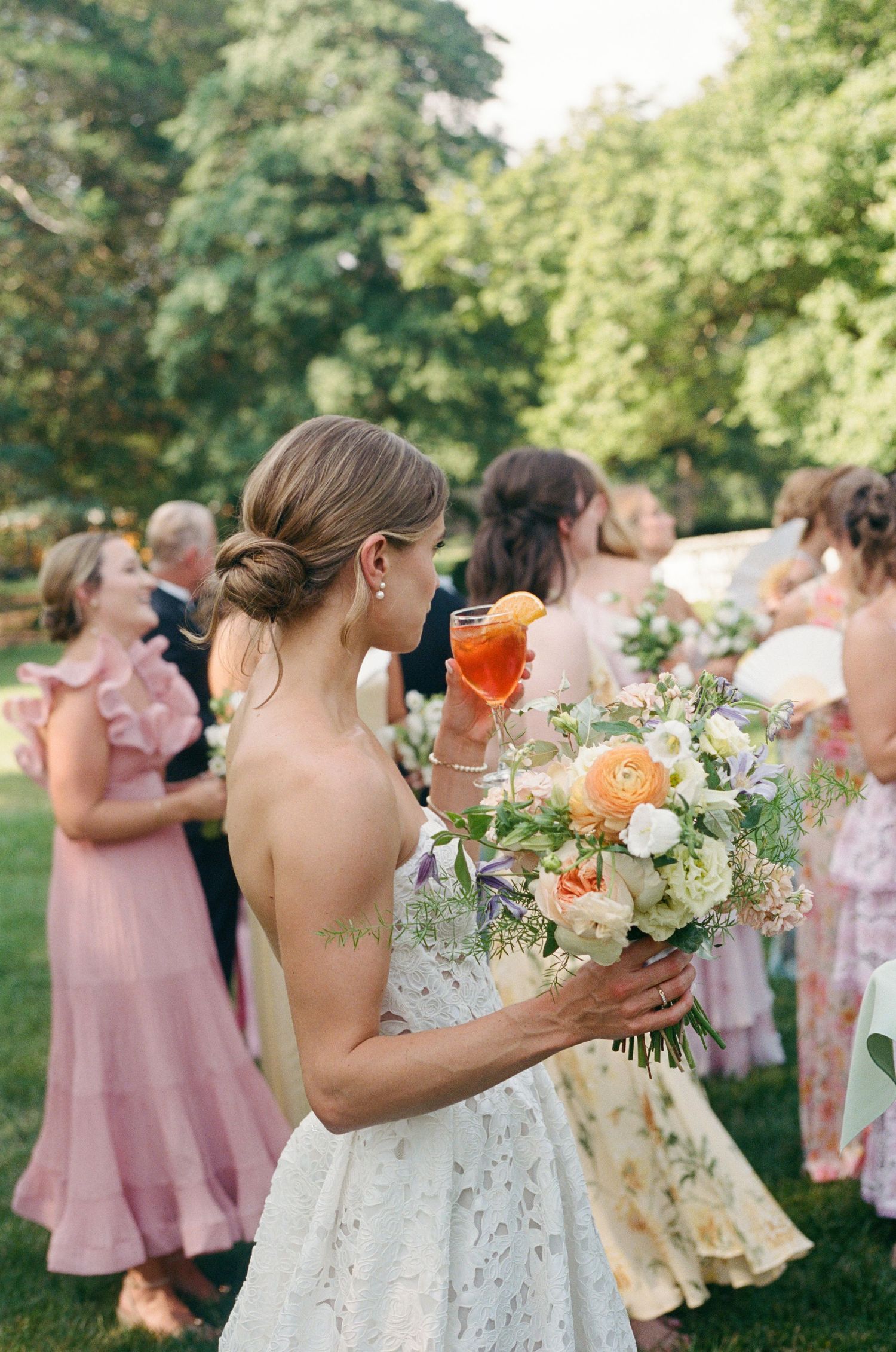 Bride holding a vibrant orange and peach bouquet surrounded by bridesmaids in pink dresses at an outdoor ceremony.
