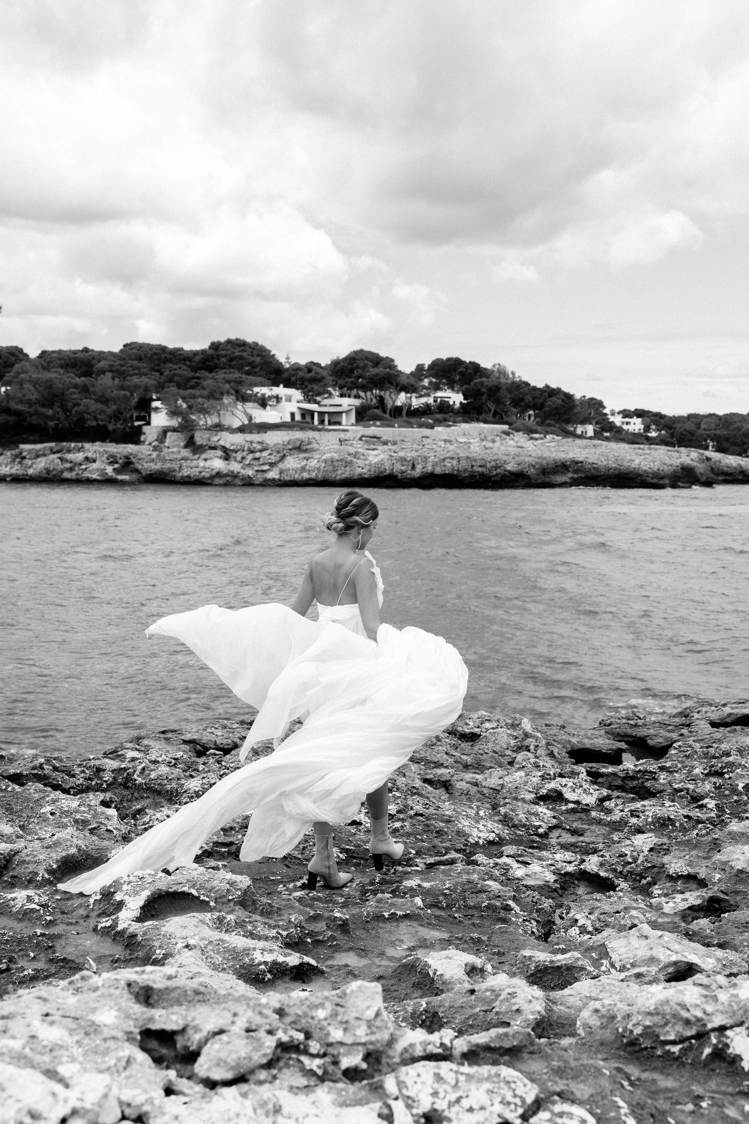 A white wedding dress flows in the wind against a rocky coastal landscape.