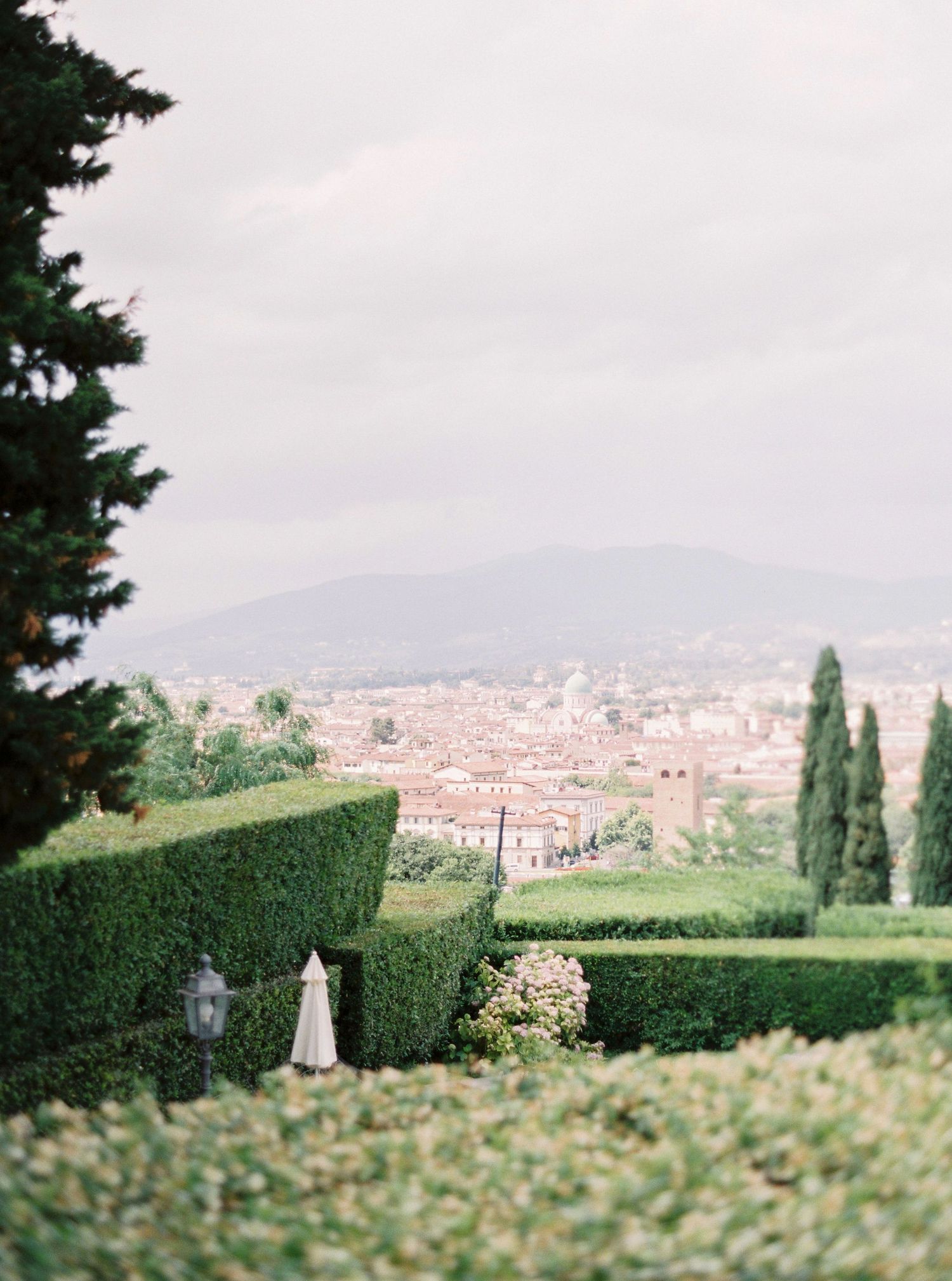 A scenic panoramic view of a European city with cypress trees and manicured hedges in the foreground.