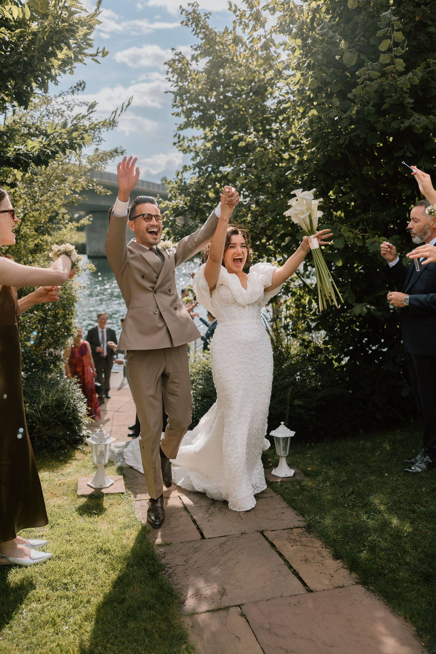 Newlyweds celebrate with guests throwing confetti on a sunny garden path.