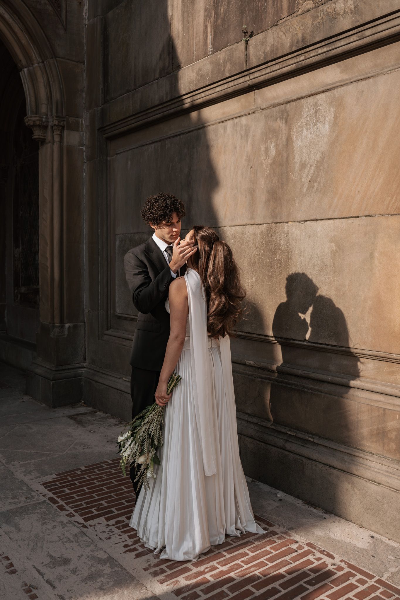 Couple in wedding attire poses against stone wall creating dramatic shadows.