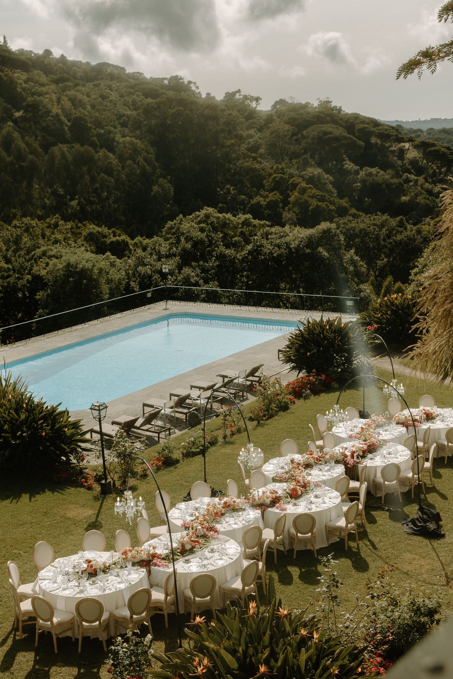 Elegant outdoor wedding reception setup with tables arranged beside a swimming pool surrounded by lush greenery.