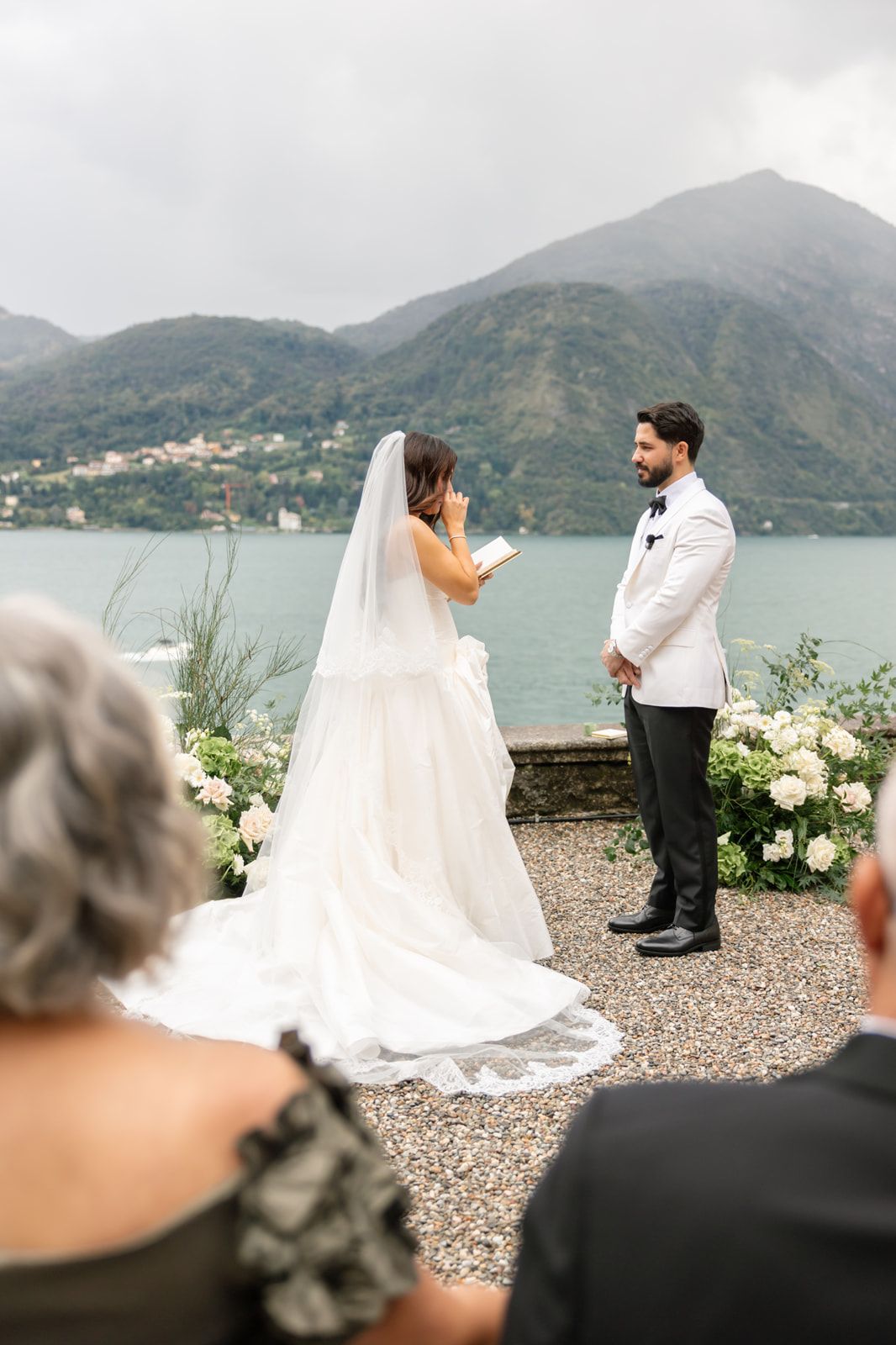 Bride in a flowing white gown reads her vows to a groom in a white tuxedo jacket during a lakeside wedding ceremony with mountain views.