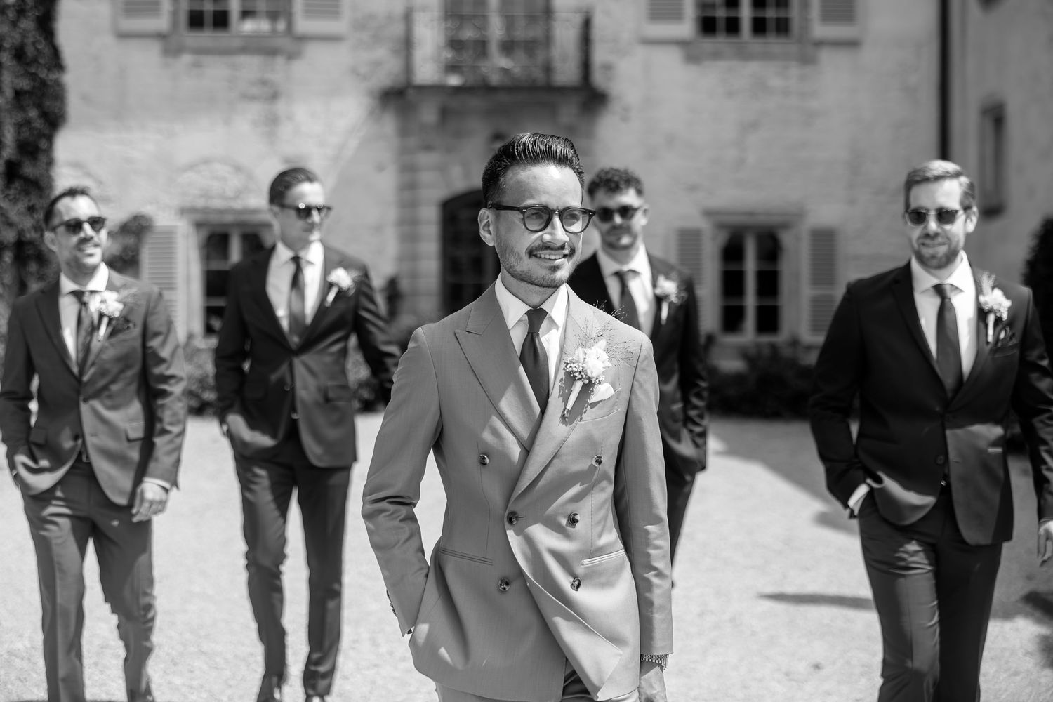 Groomsmen in suits walk together outside a grand manor house.