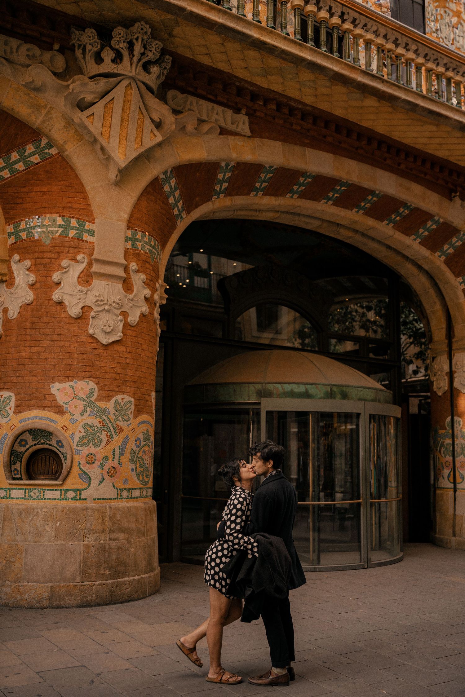 A couple shares a tender moment in an ornate architectural space with decorative arches and murals.