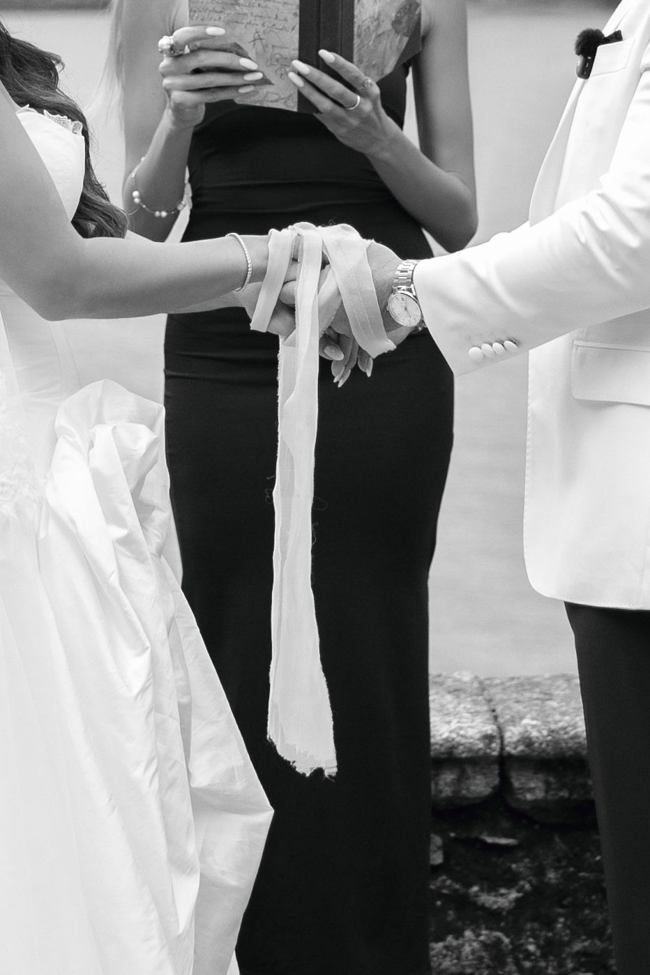 Black and white photo of a bride and groom joining hands in a wedding handfasting ceremony as an officiant in black stands behind them.