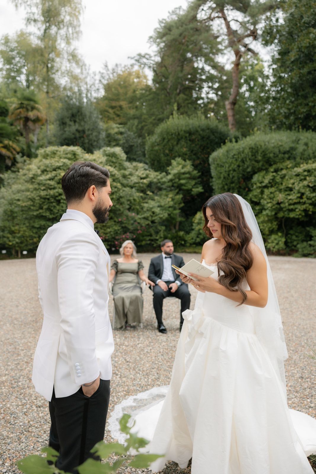 Bride in a white strapless gown and veil reads her vows to the groom in a white jacket during an outdoor garden wedding ceremony.