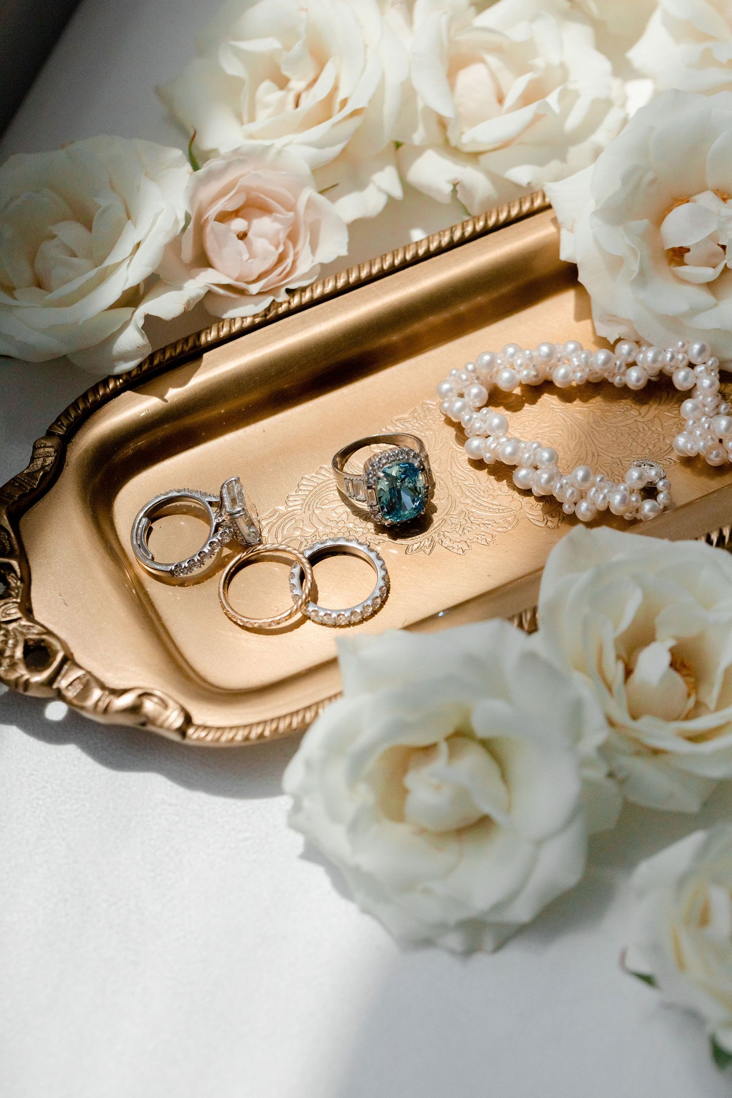 Wedding rings and pearl jewelry arranged on golden tray with white roses.