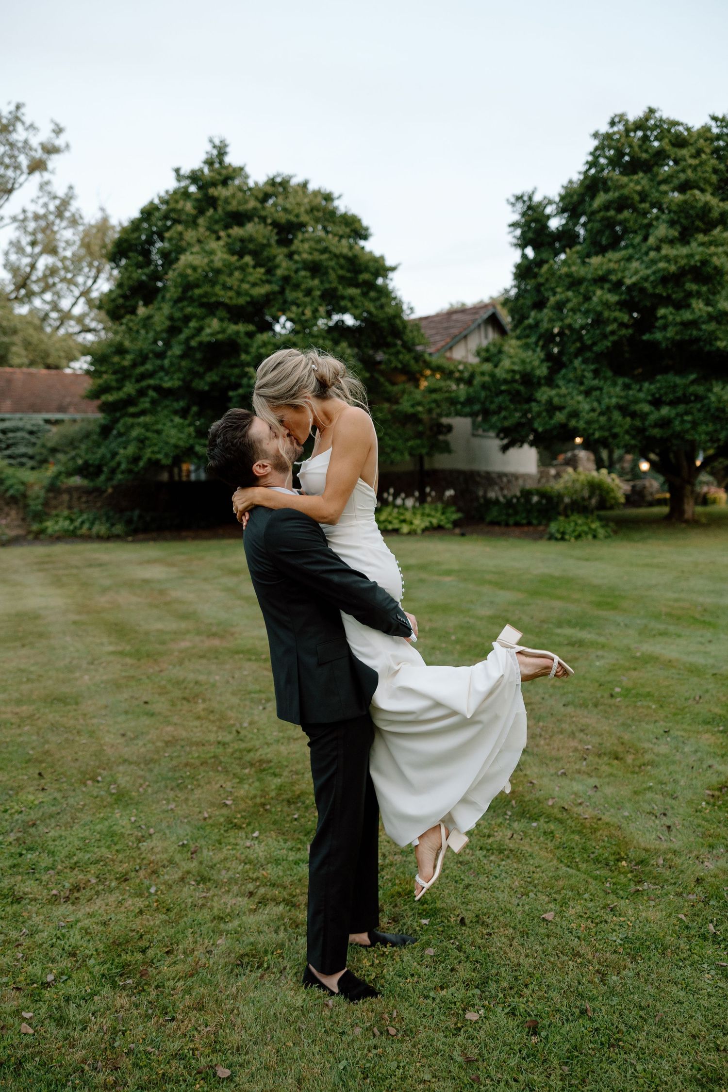 Couple shares a playful romantic moment on their wedding day in a grassy field surrounded by trees.