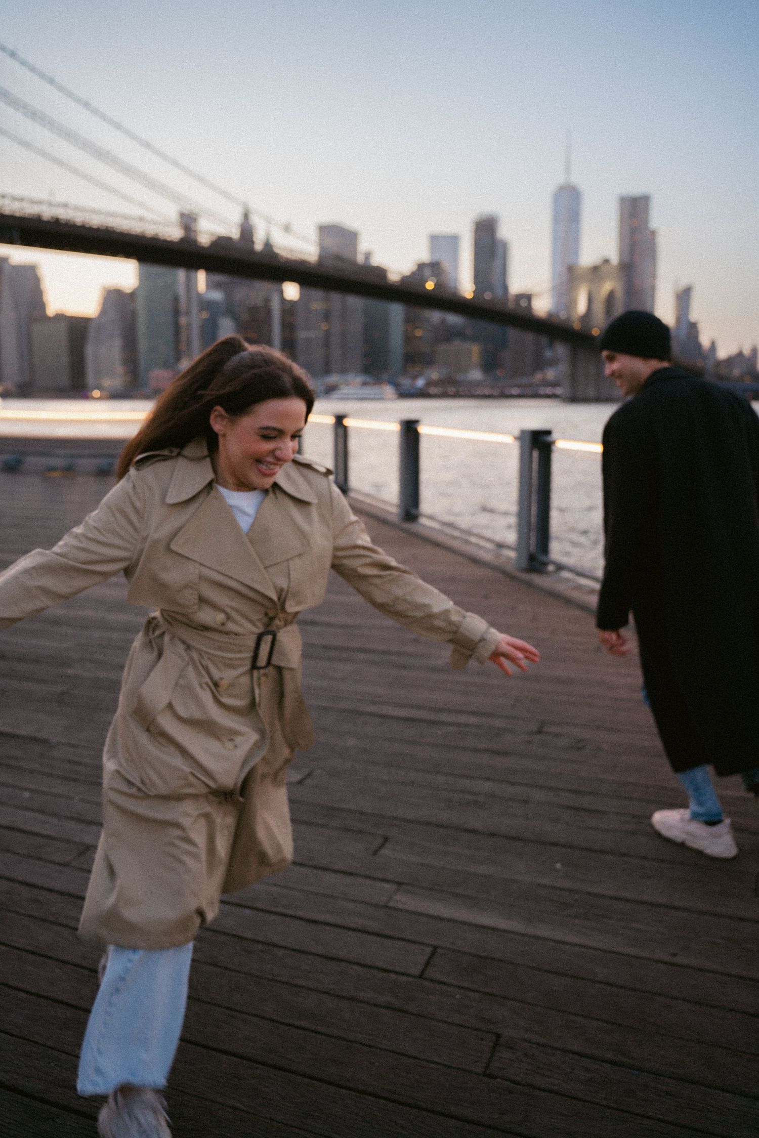Person in beige coat walking playfully along waterfront with city skyline in background.